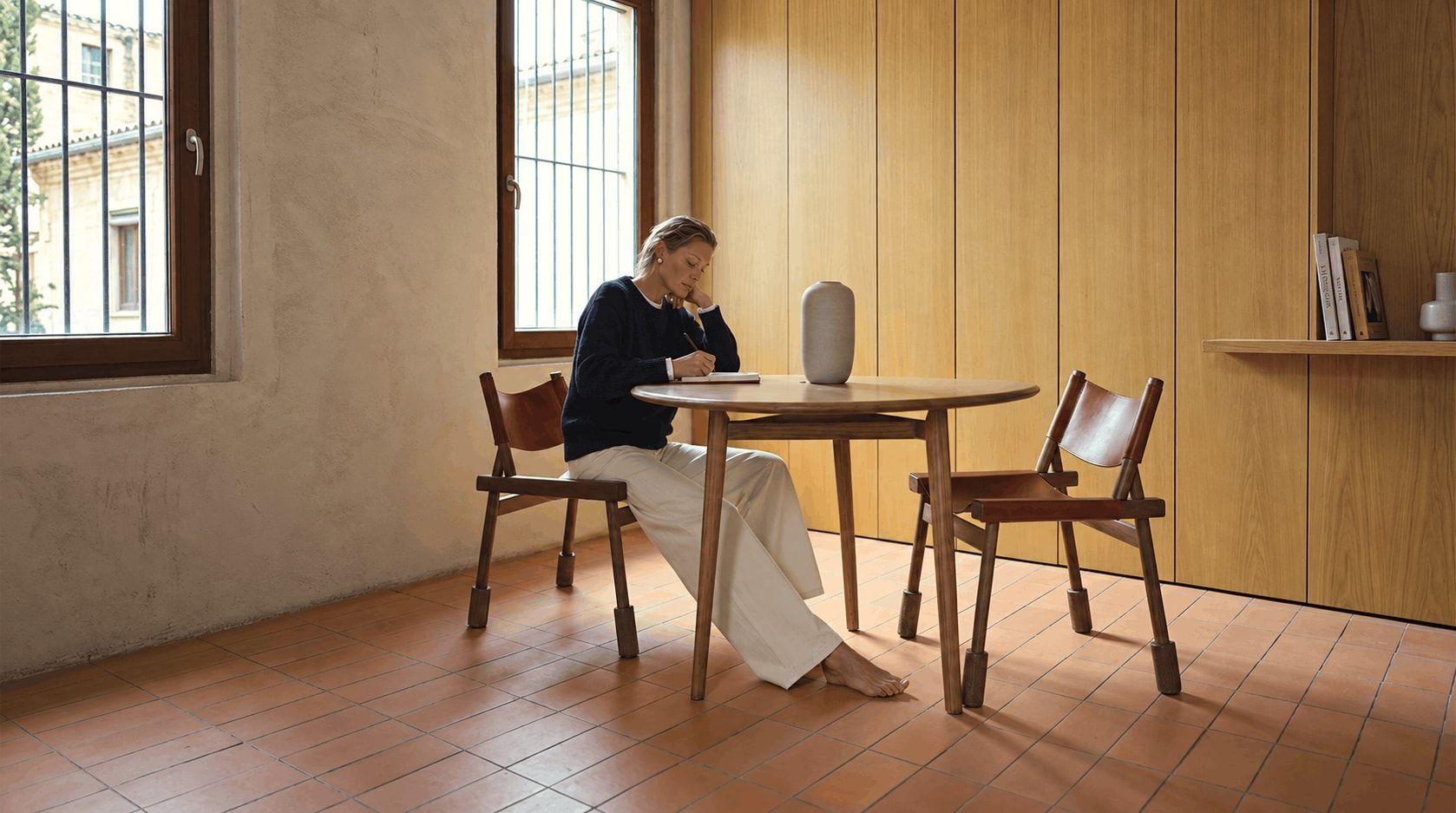 Une femme écrit dans un carnet à une table ronde en bois près d’une fenêtre, pieds nus, dans une pièce calme.